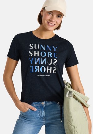 Kvinde iført beige kasket og navy T-shirt med teksten "SUNNY SHORE", holder en lysegrøn jakke og smiler til kameraet.