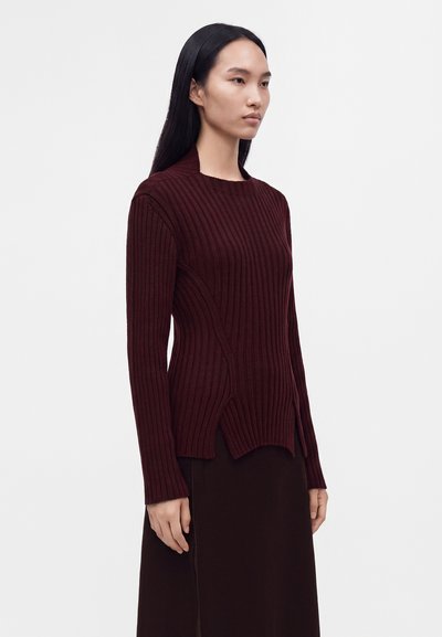 Femme aux longs cheveux noirs portant un pull côtelé bordeaux foncé et une jupe assortie, debout devant un fond clair uni.
