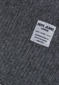 Textil gris con una textura acanalada que presenta una etiqueta tejida blanca. La etiqueta indica "Pepe Jeans London" e incluye detalles adicionales de la marca.