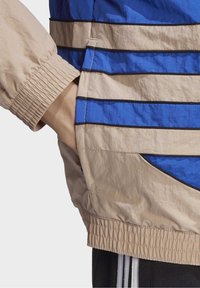 Lightweight jacket featuring horizontal stripes in blue and beige, elastic cuffs, a side pocket, and visible stitching details.