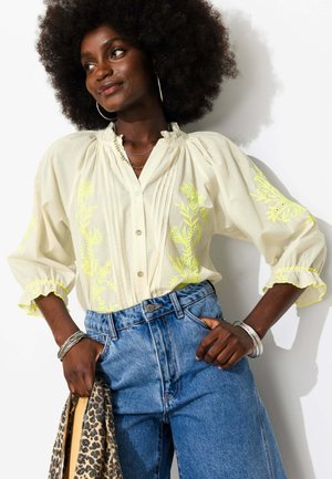 Femme avec un grand afro portant un chemisier crème avec une broderie florale jaune et un jean bleu, tenant une écharpe à imprimé léopard dans une main.