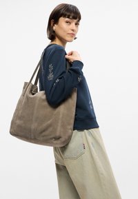 Γκρι σουέτ τσάντα ώμου με μαλακή υφή, μεγάλο σχήμα και διπλούς ιμάντες. Κεντημένα λεπτομερή σχέδια στην σκούρα μπλε μπλούζα που φορά.