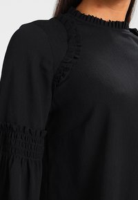 Schwarzes Langarmoberteil aus strukturiertem Stoff, mit gerafften Akzenten am Kragen und an den Schulternähten sowie Raffung an den Ärmeln.