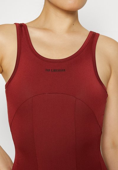 Detailný záber na osobu v priliehavom bezrukávovom vínovom tričku s nápisom "HAN KJØBENHAVN" vytlačeným na hrudi.
