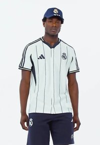 Jersey blanco a rayas con acentos en marino, con un cuello tipo V y el logo de Adidas. Lleva puesto unos pantalones cortos marinos y una gorra azul, que muestra un emblema del club.