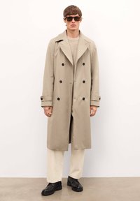 Gabardina beige con diseño de doble botonadura, botones negros y solapas amplias. Modelo combinado con un suéter crema y pantalones claros. Zapatos negros.