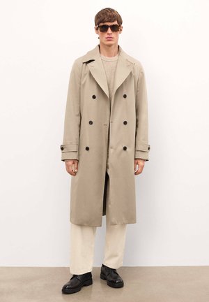Gabardina beige con diseño de doble botonadura, botones negros y solapas amplias. Modelo combinado con un suéter crema y pantalones claros. Zapatos negros.