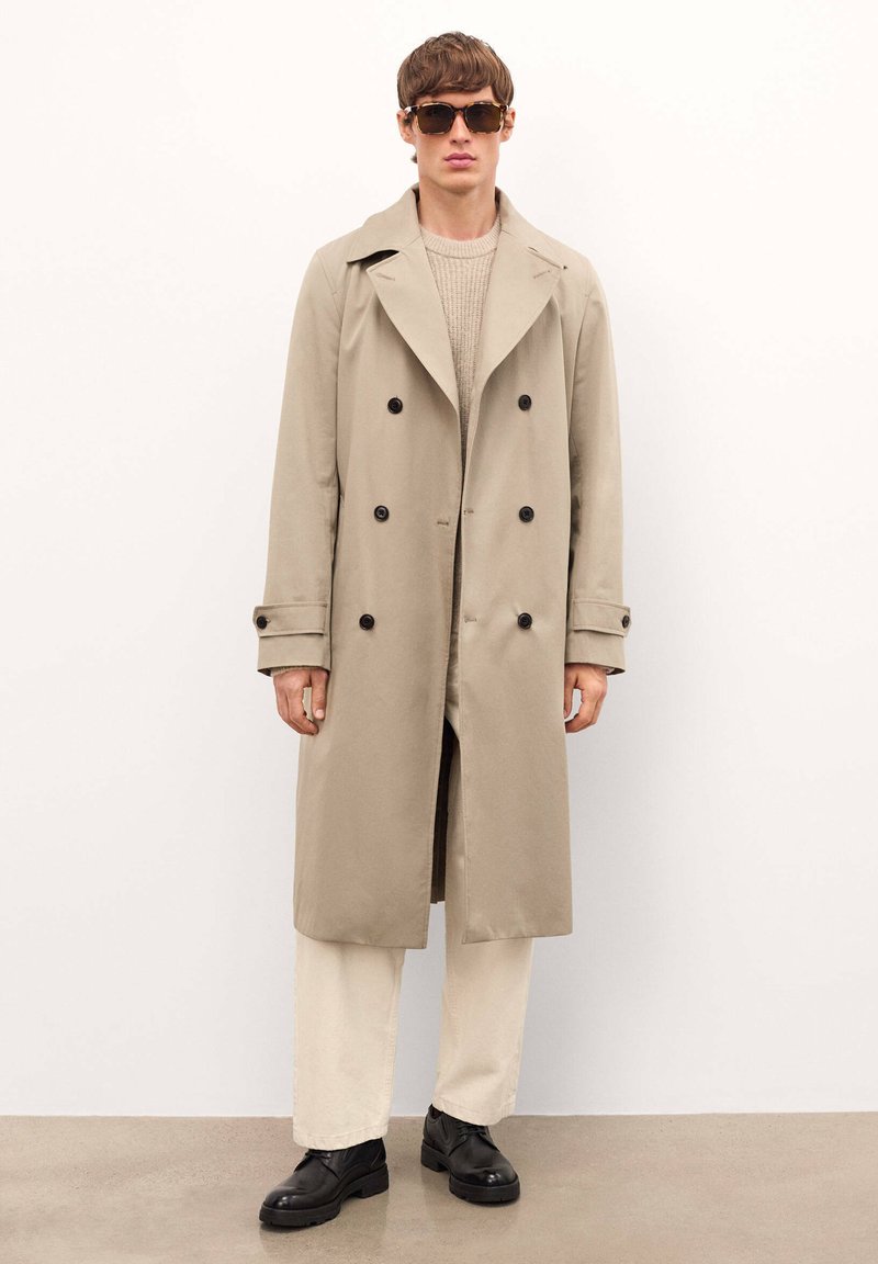 Gabardina beige con diseño de doble botonadura, botones negros y solapas amplias. Modelo combinado con un suéter crema y pantalones claros. Zapatos negros.