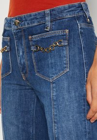 Blue denim jeans featuring two back pockets with gold chain accents, classic stitching, and a fitted design.