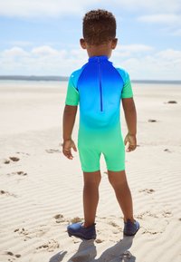 Maglia protettiva per bambini con sfumature di blu e verde a gradiente. Maniche corte, chiusura con cerniera sul retro, indossata con scarpe da acqua blu scuro. Ambientazione sulla spiaggia.