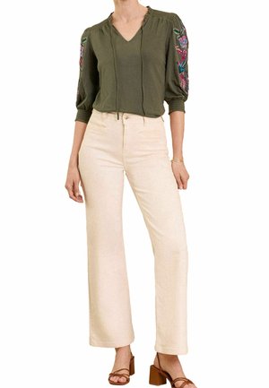 Frau trägt eine olivgrüne Bluse mit Blumenstickerei an den Ärmeln, beige, hoch taillierte, weit geschnittene Hose und braune Sandaletten mit Absatz.