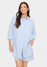 Mėlynos ir baltos juostos marškiniai su ilgomis rankovėmis, laisvas siluetas ir šoninės kišenės. Nešiojama su atitinkančiais šortais.