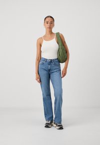 Top de alças branco com texturas, jeans azul de cintura alta, mala verde-oleosa, sapatilhas pretas e brancas, sobre um fundo neutro.