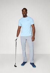 Camiseta azul clara de manga curta com um logo subtil, combinada com calças cinza claro e sapatos desportivos azuis, segurando um taco de golfe.