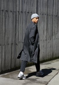 Jeune homme portant un bonnet gris et un long manteau noir marche sur le trottoir à côté d'un grand mur en bois, regardant par-dessus son épaule.