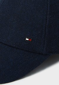Navy herringbone-patterned cap made of textured fabric, featuring a small black badge with a red, white, and blue rectangular design.