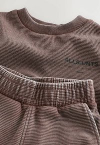 Brūns ribots džemperis ar apaļu apkakli un atbilstošas sporta bikses ar elastīgu jostu un smalku logotipa detaļu. Tekstūra ir mīksta.