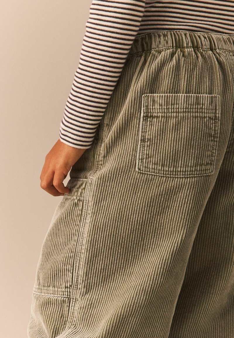 Cordhose in Olivgrün mit vertikalen Streifen, ausgestattet mit einem elastischen Bund und einer hinteren aufgesetzten Tasche. Strukturiertes Stoffdetail sichtbar.