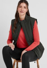 Quilted dark green vest over a red long-sleeve shirt, seated on a wooden chair. Black leggings and a casual pose complete the look.