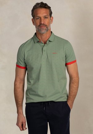 Man met grijze baard, groene polo met patroon en rode manchetten, en marineblauwe broek, staand tegen een effen achtergrond.