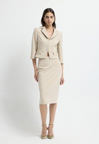 Beige taillierter Blazer mit Dreiviertelärmeln, zwei frontalen Taschen, kombiniert mit einem knielangen, figurbetonten Rock. Glattes Material mit subtiler Shed.