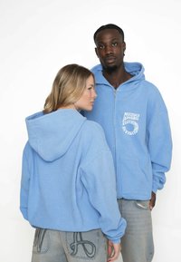 Felpe con cappuccio zip a contrasto di colore blu chiaro, in tessuto strutturato, di taglio oversize e con tasche frontali. Loghi stampati in bianco sul petto; indossate da due modelli.