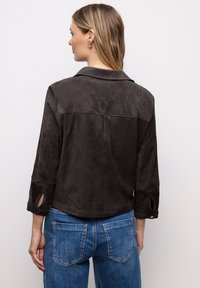 Chaqueta de ante suave en gris oscuro con cuello con solapa, mangas de tres cuartos y detalles de abertura lateral. Combinada con jeans de mezclilla.