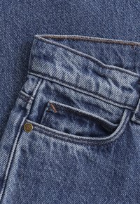 Szczegół kieszeni niebieskich dżinsów z widocznym szwem i małym metalowym nitom na tle materiału.