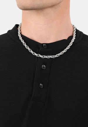 Primo piano di una persona che indossa una collana in argento su una maglietta henley nera con texture, con tre bottoni neri.