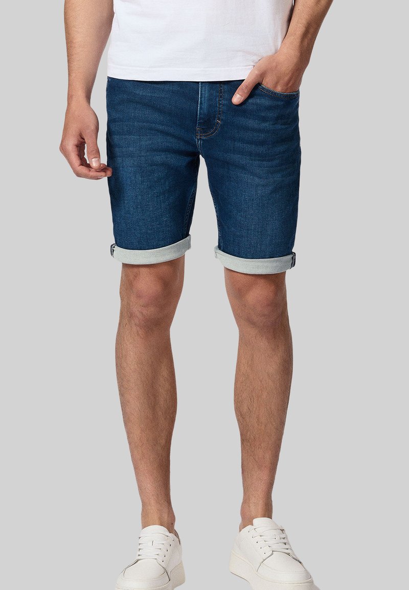 Dżinsowe szorty w ciemnoniebieskim kolorze z podwijanymi mankietami. Pięć kieszeni, widoczne szwy i efekt noszenia. Połączone z białymi sneakersami.