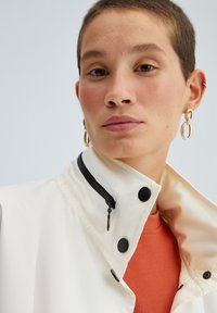 Giacca bianca con colletto alto, cerniera nera e bottoni, indossata sopra un top color ruggine-arancio. Orecchini a cerchio d'oro come accessori di rilievo.