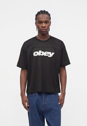 Mladý muž s pletenými vlasmi, oblečený v čiernom sieťovanom tričku s logom "obey" a modrých džínsoch, stojaci pred jednoduchým bielym pozadím.
