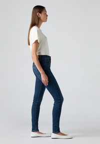 Slim fit tmavě modré džíny s světlým prošíváním, vysokým pasem, kombinované s krémovou košilí s krátkým rukávem a bílými plochými botami.