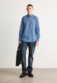 Chemise à boutons bleu à fleurs avec des manches longues, associée à un jean foncé et des chaussures noires. Le modèle tient un sac fourre-tout noir.