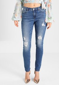 Kapeet siniset farkut, joilla on kulutusteknologisuus, jossa on repeämiä polvissa. Yhdistettynä nudenkokoisiin korkokenkiin.