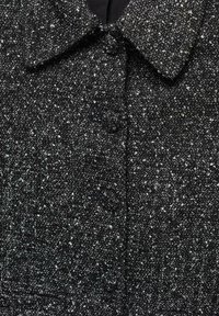 Manteau en tweed noir présentant un motif texturé, un col pointu et des boutons sur le devant. Le tissu est tissé avec des éclats blancs variés.