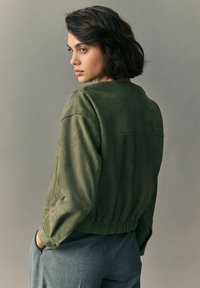 Zelená velurová bomber bunda s volným střihem, raglánovými rukávy a elastickým pasem. V kombinaci s šedými elegantními kalhotami, pohled zezadu.
