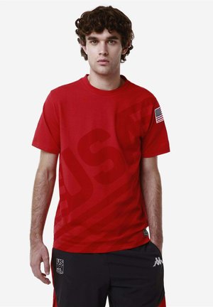 Giovane uomo che indossa una maglietta rossa con la stampa USA e una toppa con la bandiera americana, insieme a pantaloni sportivi neri con logo, in piedi su uno sfondo semplice.