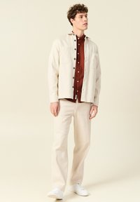 Beige button-up shirt met borstzakken, gelaagd over een roestkleurig shirt met witte knopen, gecombineerd met lichtbeige broeken en witte sneakers.