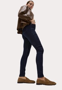 Top de cuero marrón, jeans skinny azul oscuro y mocasines de ante marrones. El atuendo cuenta con un diseño relajado y una textura suave.