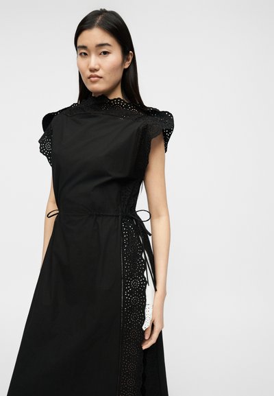 Νεαρή γυναίκα που φοράει μαύρο αμάνικο φόρεμα με λεπτομέρειες από δαντέλα και κορδόνια στο πλάι, στέκεται μπροστά σε απλό ανοιχτόχρωμο φόντο.