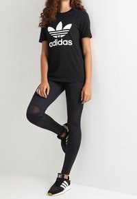 Schwarzes Adidas-T-Shirt mit weißem Logo, schwarze Leggings mit Mesh-Akzenten und schwarze Sneakers mit weißen Streifen. Das Modell posiert vor einem einfachen Hintergrund.