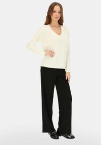 Ecru sweter z dekoltem w serek i długimi rękawami, zestawiony z czarnymi szerokimi spodniami. Modelka ma na sobie czarne buty z paskiem.
