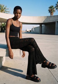 Tânără în top negru și pantaloni croșetați, așezată pe o bancă de beton în aer liber, cu palmieri și arhitectură modernă în fundal.