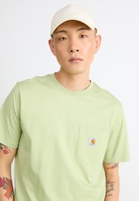 T-shirt vert clair à manches courtes en coton avec une poche poitrine et un logo Carhartt. Porté avec une casquette beige.