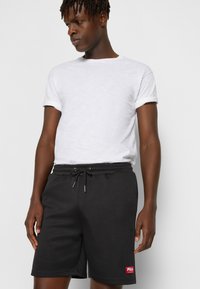 Pantaloni corti neri in felpa con cintura elastica, cordoncino e toppa rossa di Fila. T-shirt bianca a girocollo con maniche arrotolate.