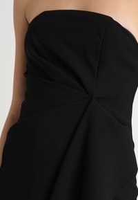 Černé šaty bez ramínek s drapovaným designem, záhyby a měkkou texturou. Tkanina je hladká, což vytváří strukturovanou siluetu.