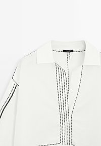 Chemise blanche avec un col classique, des détails de coutures verticales noires et une coupe décontractée avec des manches longues.