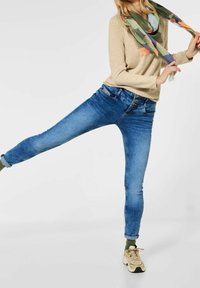 Smal blå jeans med en blekt tvätt, med framfickor och bälteöglor, kombinerat med en beige långärmad topp och en mångfärgad scarf.