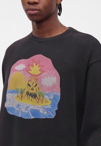 Personne portant un t-shirt noir avec un graphisme coloré d’un volcan en éruption avec un visage, des palmiers, des vagues de l’océan et un soleil couchant.